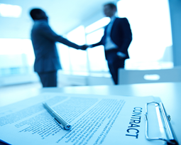 An out of focus view of two men shaking hands while a paper that says "contract" lays on the table in front of them in focus.