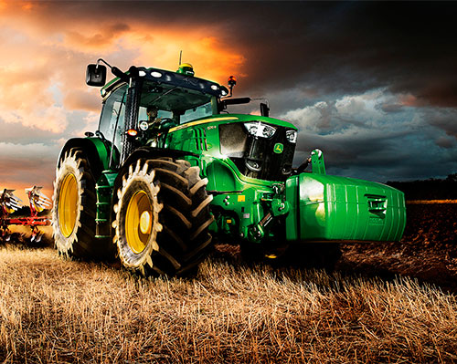 A commercial John Deere tractor with its lights on continues work while dark storm clouds loom overheard at dusk.